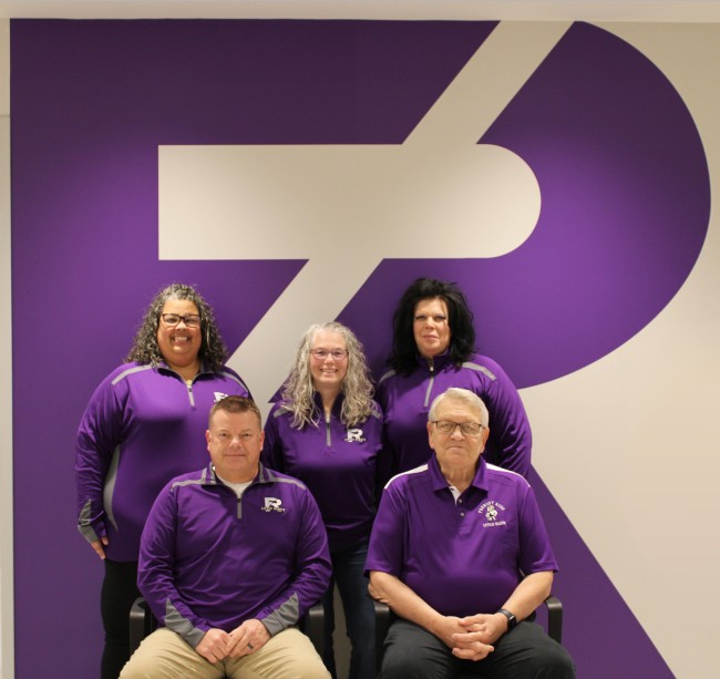 The Board of Education team posing together. The three women and two men are dressed in matching purple spirit wear, seated and standing in front of a large branded wall graphic.