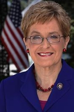 Photo of U.S. Congresswoman Marcy Kaptur wearing a blue jacket with a red necklace smiling outdoors in front of a U.S. flag