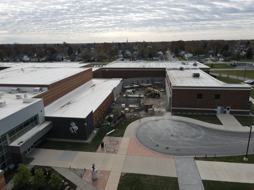 A wide-angle drone view of the entire school campus and surrounding neighborhood. The construction site for the safety connector is centrally located between two large school buildings. The foreground features a circular driveway and the main entrance, while the background shows residential streets and trees.
