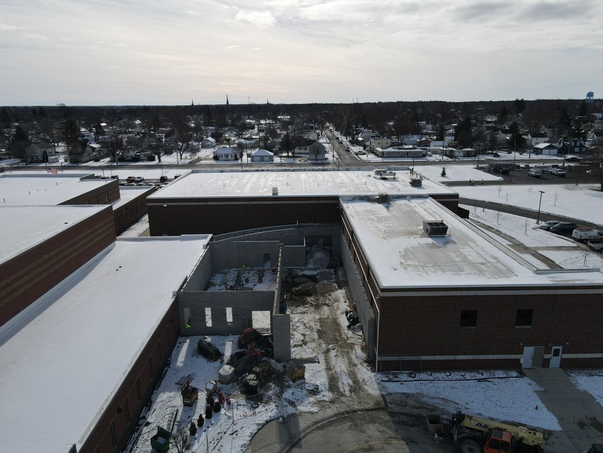 An angled aerial drone shot capturing the front of the school complex under a cloudy sky. The image shows the gap between the red brick buildings where the new connector is being built.