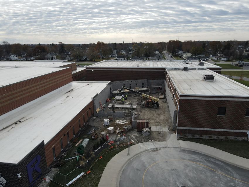 An angled aerial drone shot capturing the front of the school complex under a cloudy sky. The image shows the gap between the red brick buildings where the new connector is being built. A yellow construction lift is visible inside the work zone, and the school's mascot logo is visible on a dark wall to the left.
