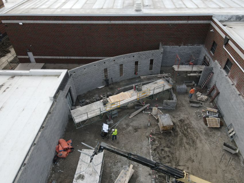 A close-up aerial view looking down into the active construction site. Gray cinder block walls are being erected, with scaffolding set up along the perimeter. Construction workers in high-visibility vests are present on the ground near pallets of materials and a telehandler boom.
