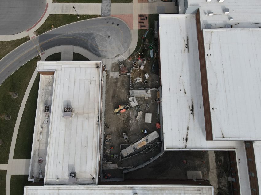 High-angle aerial drone view showing the construction footprint nestled between the high school and natatorium. To the left, a curved paved drop-off loop and sidewalks connect to the existing structures. The central construction area is active with machinery and scattered materials.