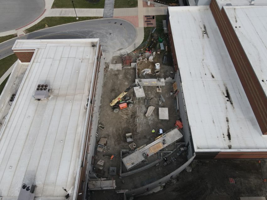 An overhead drone view looking down at the construction progress. An orange telehandler sits on the dirt ground amidst stacks of concrete blocks and building materials. The image clearly defines the workspace between the two existing white-roofed structures.