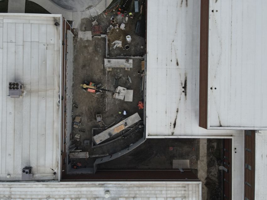 A direct overhead drone shot of the construction site located between two school buildings with white flat roofs. The narrow gap is filled with dirt, construction materials, and an orange telehandler, showing the early stages of the connector's foundation and wall assembly.